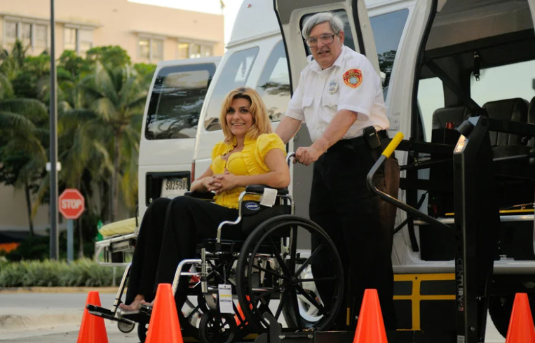 Person using a wheelchair being assisted down a wheelchair-accessible ramp from a transportation vehicle.